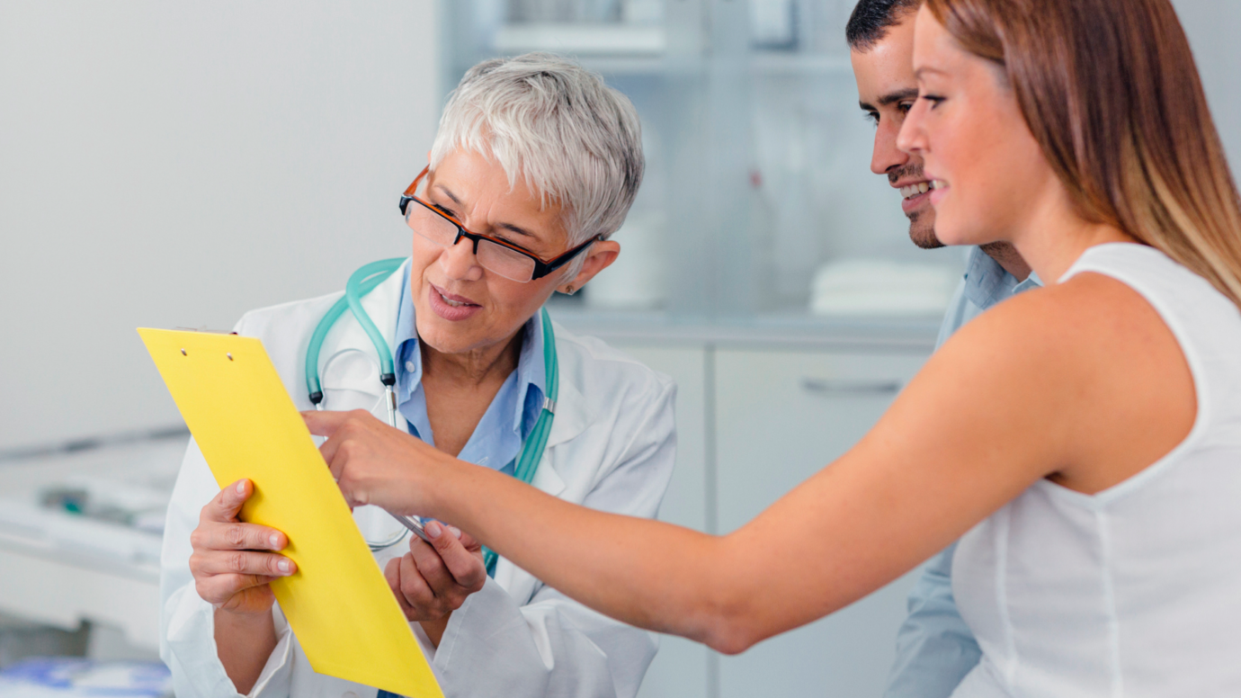 young couple pointing at clipboard being held by doctor