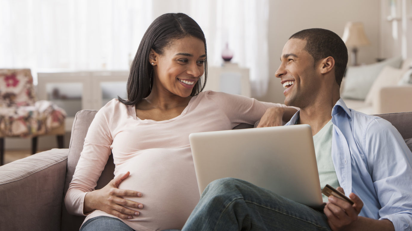 young pregnate black couple smiling on couch as women holds her belly
